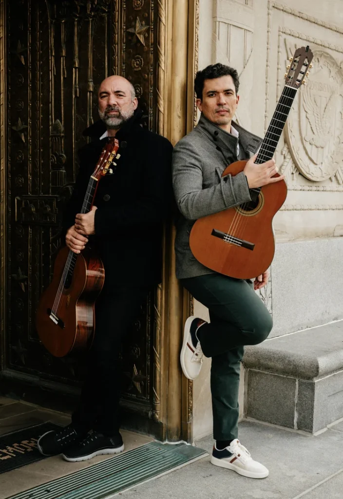 Na imagem, vemos duas pessoas lado a lado, cada uma segurando um violão acústico clássico, posicionadas diante de uma entrada arquitetônica ornamentada. O cenário inclui uma porta metálica decorada com estrelas e uma parede de pedra esculpida com um brasão, sugerindo um ambiente histórico ou artístico. Ambos estão vestidos em estilo social casual — um com casaco escuro e o outro com jaqueta cinza e calça verde — transmitindo a ideia de um contexto formal ou cultural, possivelmente ligado à música ou à fotografia artística.