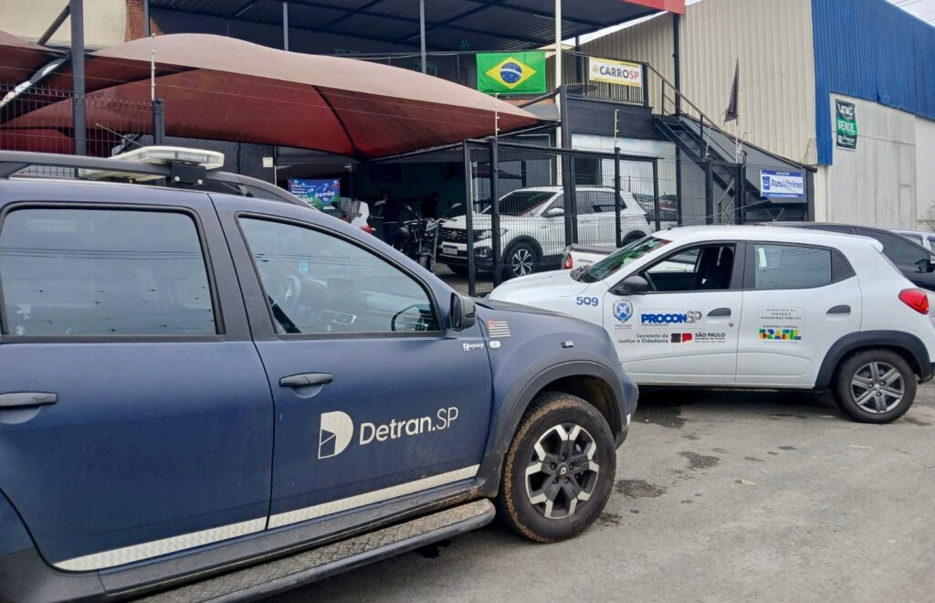 A foto mostra uma ação de fiscalização em frente a uma loja de veículos, com carros oficiais estacionados.Detalhes da cena:Primeiro plano: Esquerda: SUV azul escuro do "Detran.SP" com o logo na porta e giroflex no teto.Direita: Carro hatch branco do "PROCON-SP" nº 509. Tem adesivos da "Secretaria da Justiça e Cidadania", "Governo do Estado de São Paulo" e "Ministério da Justiça e Segurança Pública - Brasil".Fundo: Uma loja de carros chamada "CARROSP". Tem bandeira do Brasil, grade preta, toldo marrom e carros à venda no pátio. Também aparecem placas de "Auto Vitrines" e "UNIC VENDE" no prédio ao lado.Ambiente: Pátio de terra/concreto em frente ao comércio. Dia nublado.A imagem sugere uma operação conjunta entre Detran.SP e Procon-SP para fiscalização de comércio de veículos usados, algo comum pra coibir irregularidades na documentação e venda.