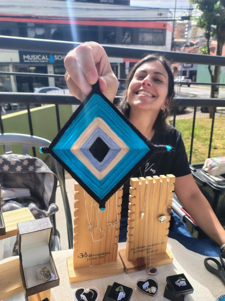 A imagem mostra uma mulher sorridente segurando um artesanato conhecido como "Ojo de Dios" (Olho de Deus), que é uma peça em formato de losango feita com fios coloridos entrelaçados. O artesanato possui camadas concêntricas de fios nas cores preta, azul clara e azul vibrante. A mulher está ao ar livre, sentada atrás de uma mesa onde há diversos itens de joalheria expostos, como anéis dentro de caixinhas, duas peças de madeira usadas para exibir os produtos e uma tesoura. Nos suportes de madeira estão impressos o nome "Consignado Joias", um número de telefone e o perfil do Instagram "@consignadojoias". Ao fundo, vê-se uma rua com algumas construções, incluindo uma escola de música, e uma grade metálica. A iluminação é intensa, típica de um dia ensolarado, e há um efeito de brilho no topo da imagem. A mulher veste uma camiseta preta e parece estar trabalhando como vendedora em uma feira ou mercado ao ar livre.