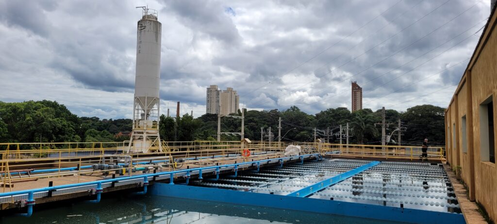 Esta imagem panorâmica mostra uma estação de tratamento de água, com um céu nublado ao fundo. A cena principal é uma estação de tratamento de água com estruturas industriais claras e complexas. Em destaque, um longo tanque de decantação ou filtração preenche a metade inferior e direita da imagem. Este tanque é parcialmente preenchido com água suja ou turva, refletindo o céu nublado acima. Sobre a água, há uma estrutura de plástico ou metal disposta em um padrão repetitivo de "ondas" ou "colmeia", projetada para aumentar a área de superfície para a filtração. Uma parte deste tanque é delimitada por uma estrutura azul brilhante. No centro da imagem, estende-se uma passarela elevada com corrimãos amarelos vibrantes, sustentada por pilares. Esta passarela é ladeada por uma intrincada rede de tubulações azuis e cinzas, algumas com válvulas e conexões visíveis, indicando o fluxo de fluidos pelo complexo. Há também um pequeno objeto enrolado em plástico branco e alguns equipamentos industriais dispostos ao longo da passarela. À esquerda da passarela, uma estrutura alta e cilíndrica, um silo branco com o logotipo "SEMAE" escrito verticalmente em azul, domina a paisagem. A superfície do silo parece um pouco manchada e desgastada pelo tempo. Mais adiante, em meio a árvores frondosas e verdes, erguem-se prédios altos e modernos com janelas retangulares e fachadas claras, contrastando com a natureza circundante. Ao fundo, um prédio de tijolos vermelhos mais fino e alto também é visível. Vários cabos elétricos e postes de transmissão cruzam o céu, adicionando outro elemento à infraestrutura industrial e urbana. No extremo direito da imagem, uma edificação baixa e retangular com uma fachada de cor ocre parece ser um anexo da estação. Há uma única pessoa visível na imagem, um homem na passarela perto da edificação ocre, vestindo roupas escuras, possivelmente um uniforme de trabalho. Ele parece estar andando e observando a área. A atmosfera geral é de um dia industrial e funcional, com o céu nublado contribuindo para uma sensação um pouco sombria, mas a presença do verde das árvores e a organização da infraestrutura sugerem um ambiente controlado e operacional.