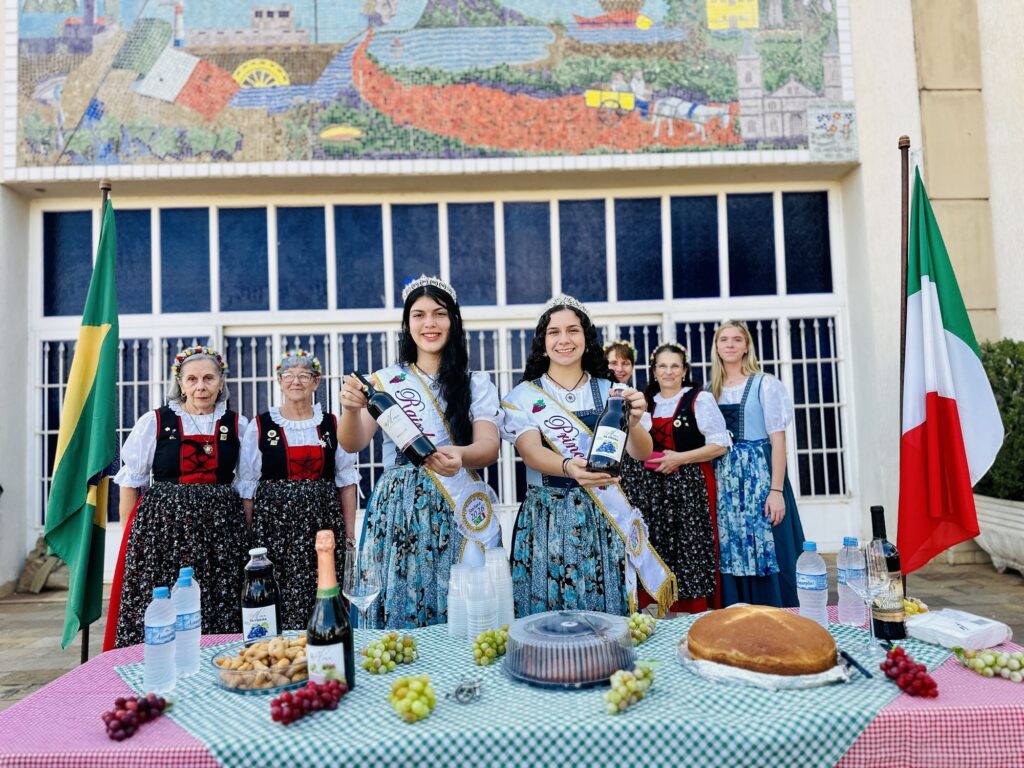 A imagem exibe um grupo de sete mulheres posando ao ar livre, em frente a uma parede decorada com um mosaico colorido e grandes janelas. Elas estão organizadas em torno de uma mesa posta com toalha xadrez verde e branca, que contém garrafas de vinho, uvas, pão e copos plásticos. * As duas mulheres centrais, que se destacam por usarem coroas e faixas, estão segurando garrafas de vinho para a câmera. * Elas vestem trajes típicos que remetem à cultura italiana, com saias floridas e aventais. * As outras cinco mulheres, que variam de idades mais maduras a jovens, também vestem trajes tradicionais semelhantes, compondo uma cena que celebra a cultura e a produção vitivinícola. * Nas extremidades da cena, há duas bandeiras hasteadas: a do Brasil à esquerda e a da Itália à direita, reforçando a temática da imigração e tradição italiana no país.