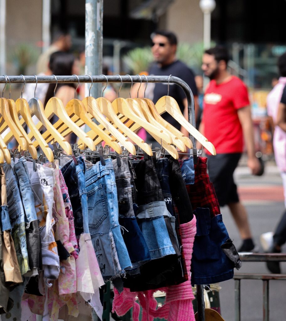 Esta foto mostra uma arara de roupas, focada em uma variedade de shorts jeans e de tecidos mistos pendurados em cabides de madeira. As peças: Os shorts são o destaque principal e exibem estilos bem variados: alguns têm visual desgastado (jeans destroyed), outros têm texturas ou padrões diferentes, incluindo um detalhe em xadrez vermelho e toques de tecidos coloridos ou desfiados. O cenário: A arara está posicionada ao ar livre ou em um espaço público, possivelmente uma feira ou mercado de rua. O fundo: O fundo está propositalmente desfocado, onde se pode ver pessoas caminhando, incluindo um homem de camiseta vermelha passando, o que sugere um ambiente movimentado e casual.