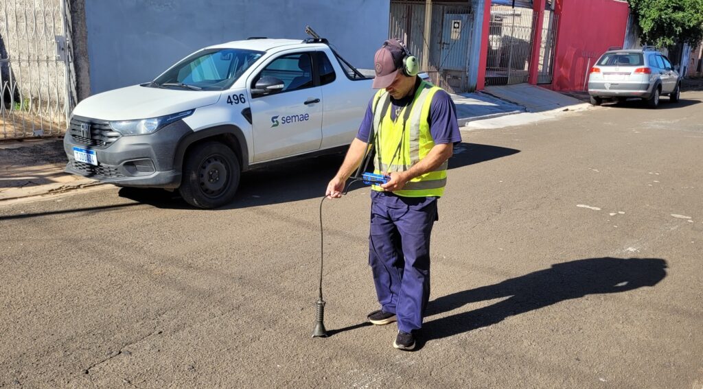 Esta imagem mostra um funcionário trabalhando em uma rua residencial. Ação: O homem, posicionado no centro, está usando um equipamento de detecção acústica ou eletrônica para verificar algo abaixo do asfalto, possivelmente em busca de vazamentos de água. Ele usa um fone de ouvido, um colete refletivo amarelo sobre uma camiseta azul marinho e calças da mesma cor, e está concentrado em um pequeno aparelho que segura com as mãos, enquanto a haste sensora toca o chão. Veículo: Atrás dele, há uma picape Fiat Strada branca, identificada com o logotipo "semae" (Serviço Municipal de Água e Esgoto) na lateral e o número "496". Ambiente: A cena acontece em uma rua pavimentada, em um dia ensolarado, o que projeta sombras nítidas no asfalto. Ao fundo, vê-se um portão metálico, paredes claras e uma casa com parede pintada de vermelho intenso. Outro carro, um modelo hatchback prateado (parece um Volkswagen), está estacionado um pouco mais adiante na rua. Detalhes: A iluminação é de luz do dia forte. O trabalhador usa um boné marrom e tênis pretos. O cenário transmite um dia típico de manutenção urbana.