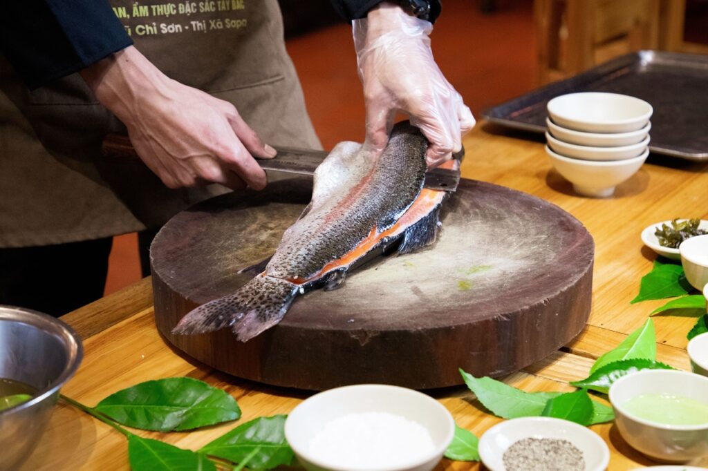 A imagem mostra uma pessoa preparando um peixe sobre uma grande tábua redonda de madeira em uma bancada de cozinha. A pessoa está usando uma luva descartável em uma das mãos e segura uma faca grande para cortar ou filetar o peixe. O peixe possui pele prateada com pequenas manchas escuras e uma faixa rosada visível ao longo do corpo, sugerindo que está sendo aberto para a retirada dos filés. Ao redor da tábua há vários recipientes brancos com temperos e ingredientes, além de folhas verdes espalhadas sobre a mesa de madeira, dando a impressão de uma preparação culinária organizada. O ambiente parece ser uma cozinha profissional ou um restaurante, com utensílios e bandejas ao fundo. A iluminação é quente e destaca os detalhes do peixe, das mãos do cozinheiro e dos ingredientes dispostos na mesa.