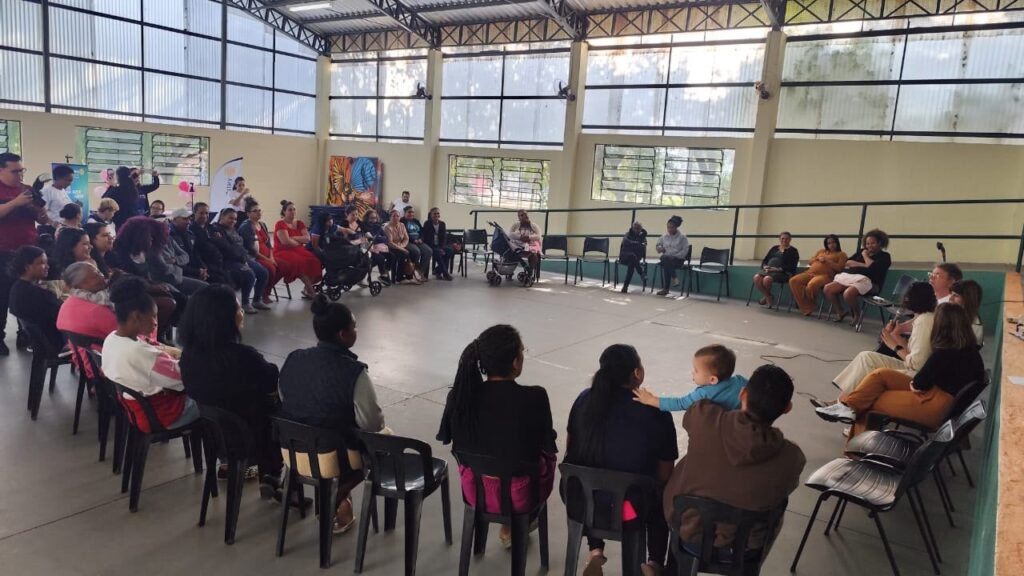 A imagem mostra um grupo de pessoas, predominantemente mulheres, reunidas em um amplo salão coberto, organizado em um formato de círculo para uma atividade ou palestra. * O ambiente parece ser uma quadra esportiva ou centro comunitário, com piso de cimento, janelas altas e estrutura metálica no teto. * As pessoas estão sentadas em cadeiras de plástico pretas, voltadas para o centro do círculo. Algumas participantes estão com crianças pequenas no colo. * À direita, um pequeno grupo parece conduzir a apresentação ou conversa, com uma pessoa segurando um microfone. * Há uma atmosfera de encontro comunitário, grupo de apoio ou reunião educativa, com uma disposição que favorece a interação e o diálogo entre todos os presentes.