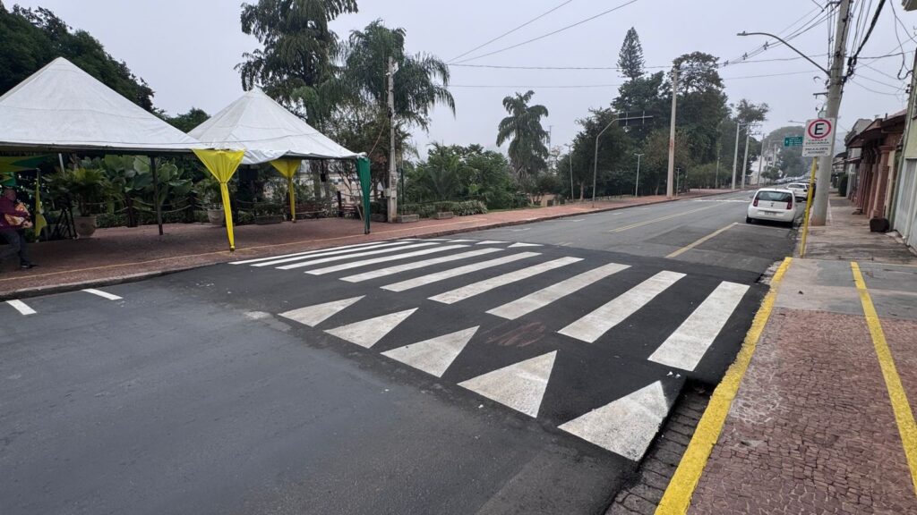 A imagem mostra uma rua com uma faixa elevada para pedestres pintada com listras brancas e triângulos, que funciona como um redutor de velocidade. Ao lado da rua, há uma calçada com pedras vermelhas e um meio-fio amarelo. Do lado esquerdo da imagem, estão montadas duas tendas brancas com detalhes amarelos, possivelmente para algum evento ou venda. Uma pessoa está próxima a uma das tendas. Na lateral direita da rua, há uma placa de trânsito indicando proibição de estacionamento. Ao fundo, há um carro branco estacionado e várias árvores verdes, sob um céu nublado. No chão, próximo à faixa elevada, está escrito em português: "E CARGA E DESCARGA PISCA ALERTA ACIONADO", o que sugere uma área destinada para carga e descarga com alerta piscante. A cena transmite um ambiente urbano tranquilo, possivelmente em um bairro residencial ou comercial pequeno.