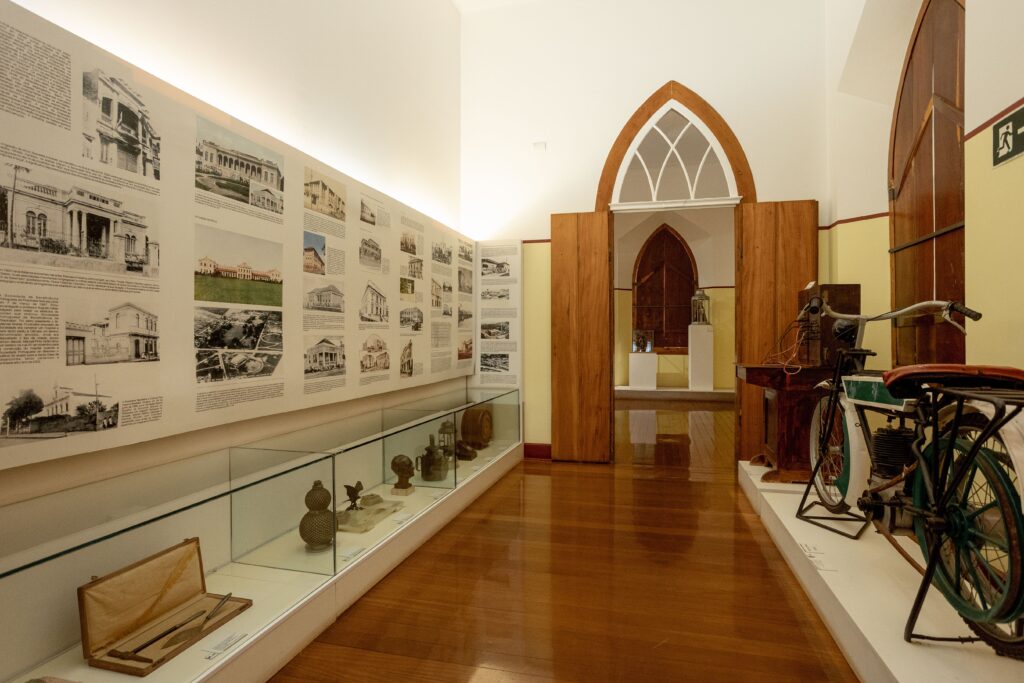 Na imagem, vemos o interior de uma sala de exposição em um museu histórico ou cultural. À esquerda, há um longo painel com fotografias antigas e textos explicativos sobre construções arquitetônicas, acompanhado de vitrines de vidro que exibem ferramentas, objetos decorativos e instrumentos antigos. À direita, sobre uma plataforma branca, destaca-se uma bicicleta vintage com componentes mecânicos, possivelmente um modelo motorizado inicial. O espaço possui piso de madeira polido, portas em arco com acabamento branco e iluminação suave que valoriza os itens expostos. A cena transmite a atmosfera de preservação histórica e patrimônio cultural, ressaltando a importância da tecnologia antiga e da arquitetura como parte da memória coletiva.