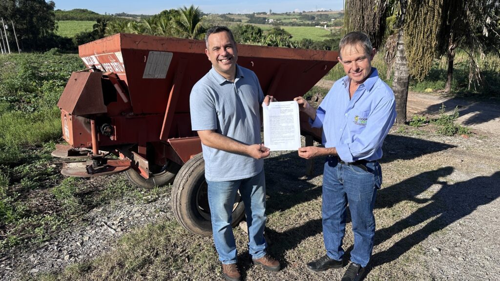Esta imagem mostra dois homens sorridentes em um ambiente rural, posando para uma foto enquanto seguram um documento. As Pessoas: À esquerda, um homem veste uma camisa polo cinza e jeans. À direita, outro homem, usando uma camisa social azul claro com o logotipo da "COOPHORT" (Cooperativa de Produtores de Hortifrutis), também de jeans. Ambos estão sorrindo para a câmera. O Documento: Eles seguram juntos, com ambas as mãos, uma folha de papel que parece ser um documento oficial ou contrato. O Cenário: Atrás deles, há um grande equipamento agrícola de cor avermelhada (parece ser um distribuidor de calcário ou adubo) estacionado em uma estrada de terra. O fundo mostra uma paisagem de campo ensolarada, com plantações e vegetação ao longe, típica de uma área agrícola.