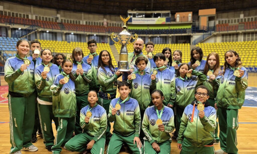 A imagem mostra uma equipe esportiva reunida dentro de um ginásio coberto, posando para uma foto de grupo. Os atletas, tanto homens quanto mulheres, estão usando uniformes combinando nas cores azul e verde, com o logo "DEDINI" visível nas roupas. Todos parecem muito felizes e orgulhosos, exibindo medalhas de ouro penduradas no pescoço. No centro do grupo, um dos integrantes segura um grande troféu dourado, simbolizando uma conquista importante. O ginásio ao fundo tem arquibancadas amarelas e um piso de madeira, indicando um ambiente profissional para competições esportivas. A foto transmite um clima de celebração e vitória da equipe.