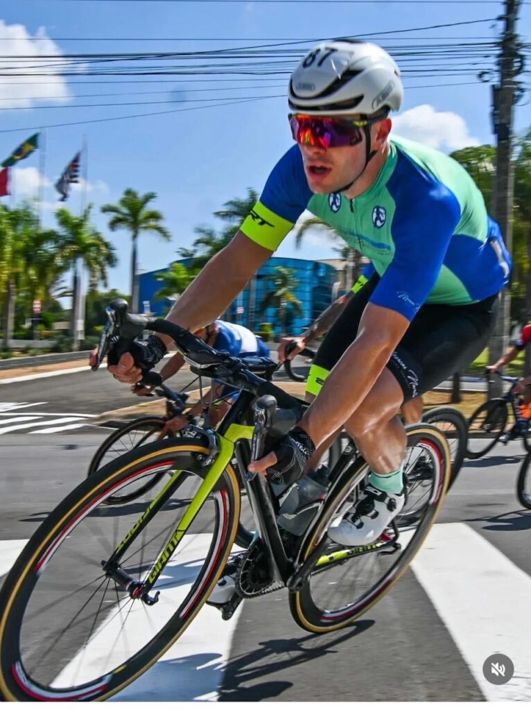 Esta imagem captura um momento de foco e esforço intenso em uma corrida de ciclismo de estrada. Destaque: Um ciclista em primeiro plano pedala concentrado, vestindo uma camisa azul e verde, shorts pretos, capacete branco (com o número 87) e óculos refletivos coloridos. Ação: Ele segura o guidão de sua bicicleta de estrada (da marca Giant) com firmeza, atravessando o que parece ser uma faixa de pedestres em uma rua pavimentada. Ambiente: É um dia ensolarado. Ao fundo, pode-se ver outros ciclistas seguindo o pelotão, além de palmeiras, bandeiras e um edifício azul, sugerindo uma competição urbana ou em rodovia movimentada.