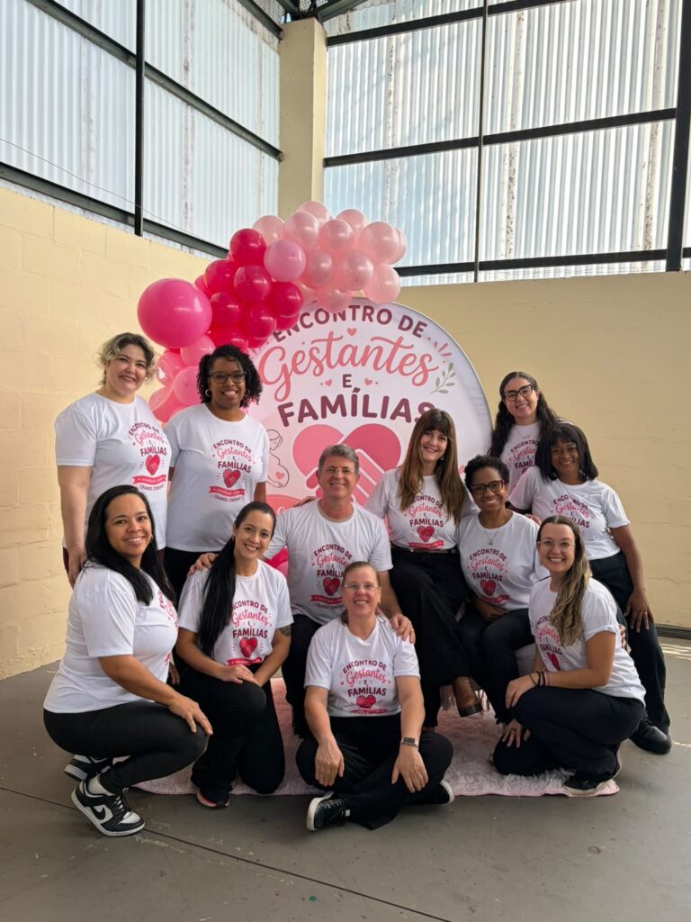 Esta imagem mostra um grupo de 11 pessoas, sendo 10 mulheres e 1 homem, posando para uma foto em um evento intitulado "Encontro de Gestantes e Famílias". * O grupo está reunido em frente a um painel circular decorado com balões cor-de-rosa de vários tons. O painel traz o nome do evento e ilustrações de mãos unidas em formato de coração. * Todos os participantes vestem camisetas brancas padronizadas com a logomarca do evento e calças pretas. A disposição é composta por algumas pessoas em pé, na parte de trás, e outras agachadas ou sentadas à frente, criando uma composição organizada. * O ambiente parece ser um espaço amplo, possivelmente um salão ou quadra, com paredes de blocos aparentes e uma estrutura de teto industrial. A atmosfera é de celebração, acolhimento e trabalho em equipe.