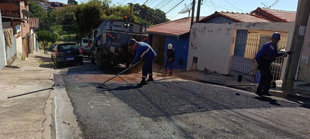 A imagem mostra uma cena de reparo de rua em uma área residencial durante o dia. Três trabalhadores, vestidos com uniformes azuis e capacetes de segurança, estão pavimentando um trecho da via com asfalto fresco. Um dos trabalhadores está usando um ancinho para espalhar o asfalto, enquanto os outros dois utilizam ferramentas para compactar e finalizar a superfície. Ao fundo, há um caminhão azul estacionado na rua, parcialmente bloqueando a visão, e um carro Volkswagen Fox preto estacionado atrás do caminhão. As casas ao redor têm telhados de cerâmica vermelha e paredes de concreto. A iluminação é forte, indicando um dia ensolarado, e as sombras são bem definidas. O asfalto fresco tem uma textura escura e rugosa, contrastando com o concreto mais claro e desgastado da estrada já existente. A cena transmite uma sensação de trabalho rotineiro de manutenção urbana.