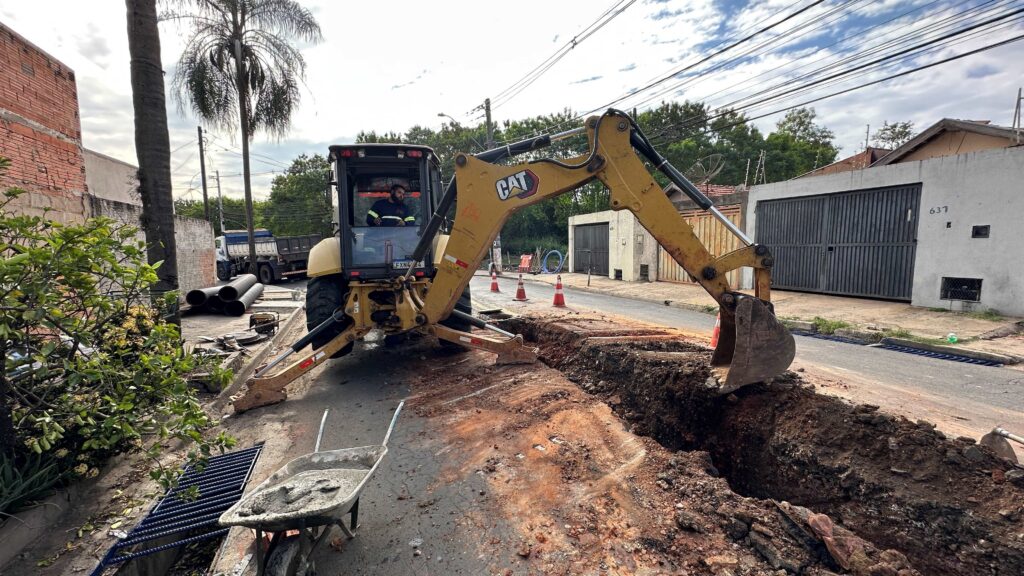 A imagem mostra uma cena de uma obra em uma rua residencial durante o dia. No centro da imagem, há uma retroescavadeira amarela da marca CAT, com um operador visível dentro da cabine, que está escavando uma vala ao lado da rua. No chão, próximo à máquina, há uma carrinho de mão com mistura de concreto e algumas tubulações pretas. Ao redor da área escavada, há cones de sinalização para garantir a segurança. Ao fundo, é possível ver casas com portões cinzas, postes de energia com cabos e um céu claro com algumas nuvens. A rua está parcialmente aberta para a obra, com terra e asfalto visíveis no local da escavação. O número 637 está pintado na parede de uma das casas ao fundo, e a placa da retroescavadeira é FXN4J84. A cena transmite a atividade típica de construção ou manutenção urbana.