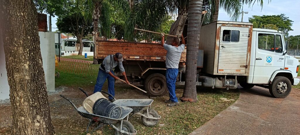 A imagem mostra uma cena ao ar livre, provavelmente em um parque ou área verde, onde dois trabalhadores estão realizando serviços de limpeza ou manutenção. Ambos vestem camisetas cinza e calças jeans. Um dos trabalhadores está usando uma pá para colocar material em um carrinho de mão, enquanto o outro está usando uma pá para carregar material na caçamba de um caminhão basculante de cor ferrugem. Ao lado deles, há um caminhão branco com a inscrição "PIRACICABA Prefeitura do Município" e um brasão, indicando que o serviço é realizado por uma prefeitura municipal. No carrinho de mão, há um balde preto e um recipiente plástico bege. Ao fundo, é possível ver árvores grandes, gramado e uma calçada, sugerindo que o local é uma área pública. A iluminação indica que a foto foi tirada durante o dia. A cena transmite uma atividade rotineira de manutenção urbana.