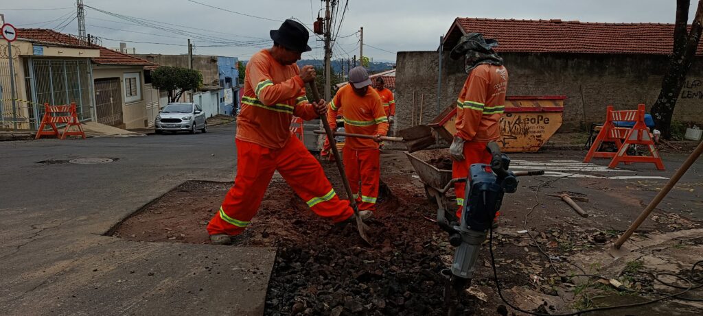 A imagem mostra uma cena de manutenção viária em um dia nublado. Três trabalhadores, todos vestidos com uniformes laranja com faixas refletivas amarelas, estão realizando reparos no asfalto da rua. Dois deles usam pás para cavar e limpar a área, enquanto o terceiro está operando uma britadeira próxima a uma carroça cheia de terra. Um deles usa um chapéu preto, outro um boné cinza, e o terceiro um chapéu largo para proteção. Ao fundo, há casas residenciais, um carro prata estacionado e um contêiner amarelo com a inscrição "LeoPe" e números de telefone. Também é possível ver barreiras laranjas de segurança ao redor da área de trabalho. O cenário transmite o esforço físico e a organização envolvidos na manutenção urbana.
