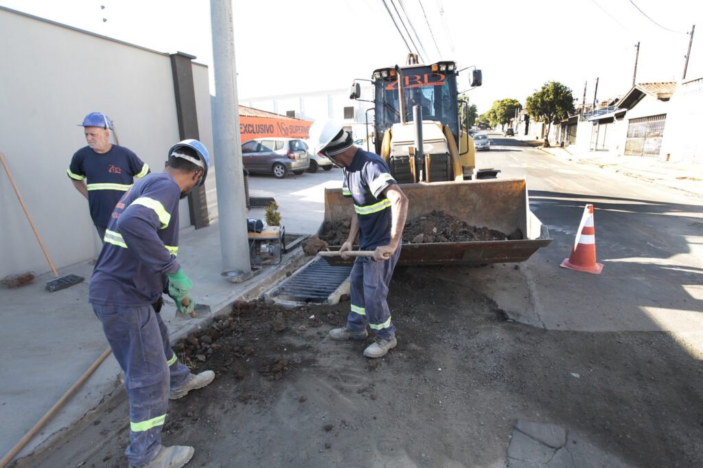 Na imagem, vemos uma cena de manutenção urbana: trabalhadores com uniformes refletivos e capacetes realizam serviços de limpeza ou reparo próximo a um bueiro, utilizando pás para remover terra ou entulho. Ao lado, uma retroescavadeira com a caçamba cheia de terra auxilia na tarefa, enquanto um cone laranja sinaliza a área de trabalho. O cenário acontece em uma rua de bairro, com casas, veículos e postes ao fundo, transmitindo a importância da infraestrutura urbana e da segurança viária para o bom funcionamento da cidade.