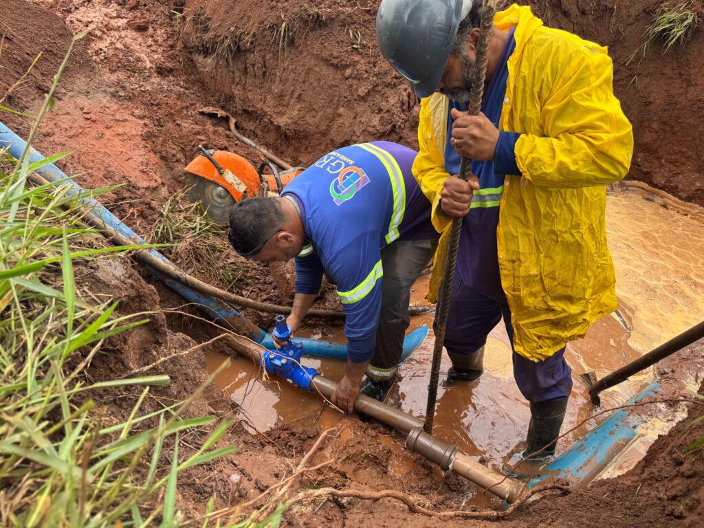 A imagem mostra dois trabalhadores em um canteiro de obras, dentro de uma vala cheia de lama e água. Um dos homens está agachado, vestindo uma camisa azul com a inscrição "DGR SANEAMENTO" e faixas refletivas, mexendo em uma tubulação de água, provavelmente fazendo algum reparo ou ajuste em uma válvula azul conectada a um cano marrom. O outro homem está em pé, usando um capacete cinza, uma capa de chuva amarela por cima de roupas escuras e botas de borracha pretas, segurando uma barra de metal vertical. O ambiente é de terra úmida e lama, com a vala cercada por paredes de terra vermelha. Ao fundo, há uma serra elétrica laranja sobre a lama. A cena transmite uma sensação de trabalho pesado em condições difíceis, possivelmente relacionado à manutenção ou instalação de infraestrutura de saneamento básico.