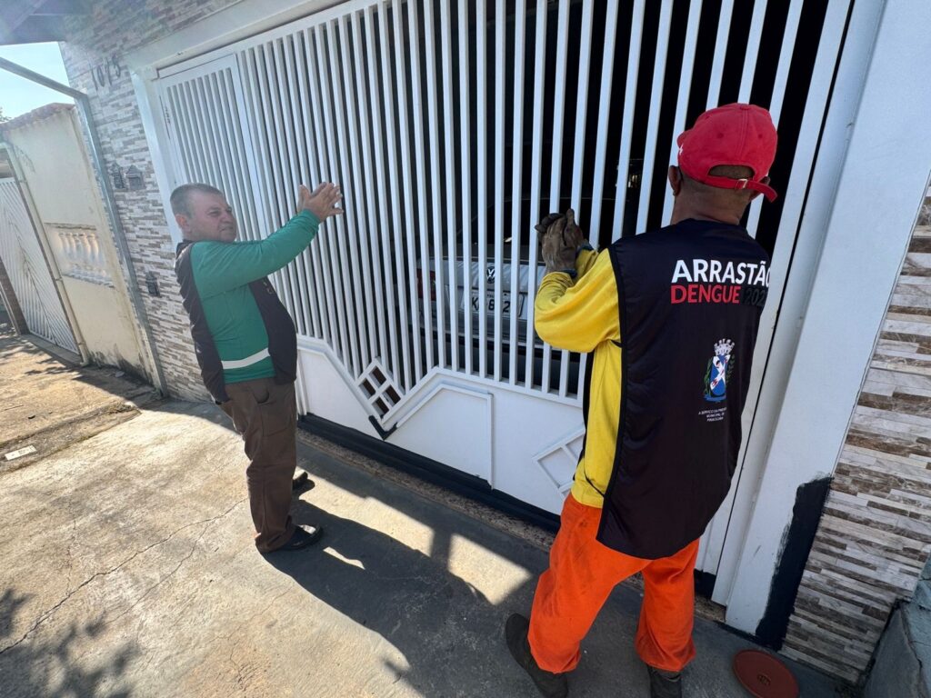A imagem mostra dois homens em frente a um portão branco de uma residência, realizando uma ação de fiscalização. * O homem à direita veste um colete preto com a inscrição "ARRASTÃO DENGUE 2023" e o brasão da Prefeitura de Piracicaba, indicando que se trata de um agente de saúde ou funcionário municipal em uma campanha de combate à dengue. * O homem à esquerda, que veste uma camisa de manga longa verde e colete, gesticula com as mãos em direção ao interior da casa, possivelmente conversando com o morador ou orientando o agente. * Atrás do portão, é possível visualizar parte da traseira de um veículo Volkswagen estacionado. O cenário é uma rua pavimentada sob luz solar intensa, o que projeta sombras nítidas dos indivíduos no chão.