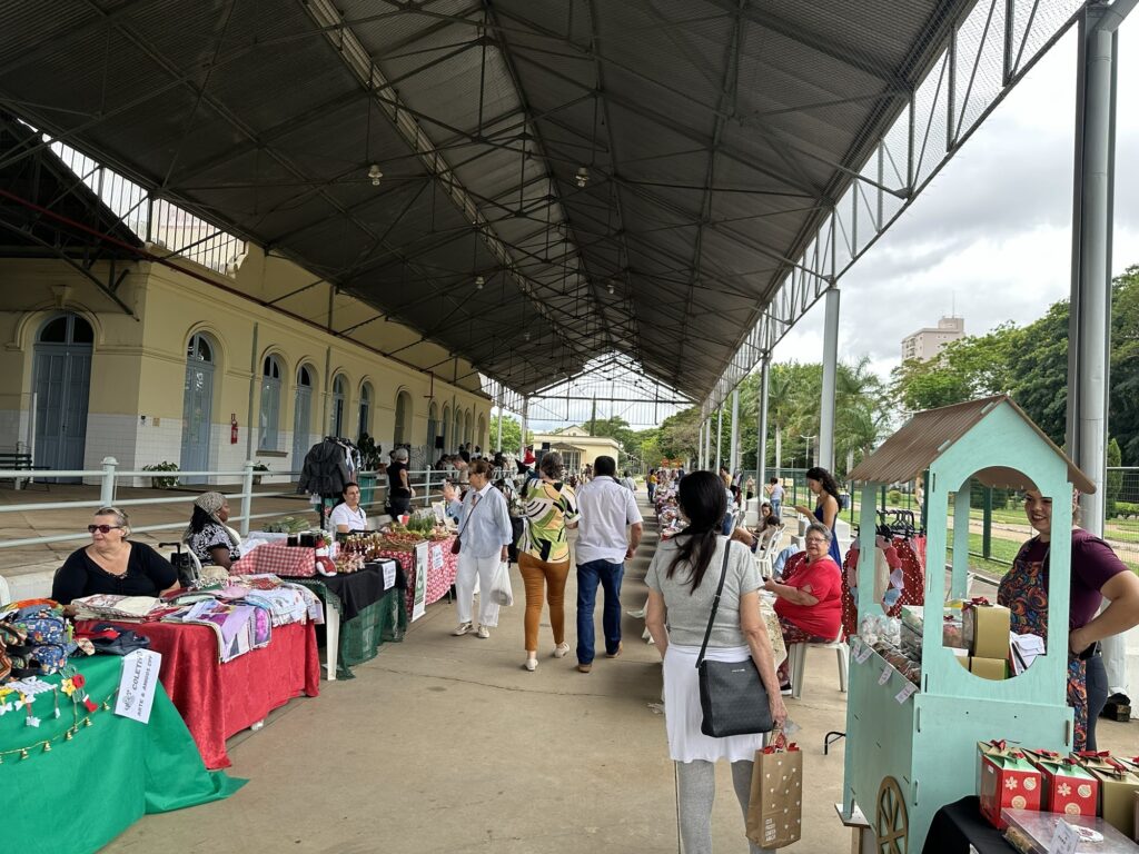 A imagem mostra um mercado de artesanato ao ar livre, situado sob uma estrutura metálica com teto alto e transparente, que permite a entrada de luz natural. À esquerda, há um prédio antigo, pintado de amarelo claro, com janelas e portas arqueadas, que parece ser parte do local onde o mercado acontece. Ao longo do prédio, várias barracas estão montadas com toalhas coloridas, exibindo diferentes produtos artesanais, como tecidos, potes e outros objetos feitos à mão. No centro da imagem, uma mulher com cabelo escuro, vestindo uma blusa cinza e uma saia branca, caminha de costas, carregando uma bolsa preta e uma sacola de compras. À direita, há uma espécie de carrinho ou estande azul claro em forma de casinha, onde outra mulher, vestida com uma blusa colorida, está atendendo ou organizando os produtos expostos. Ao fundo, é possível ver árvores e alguns edifícios, sugerindo que o mercado está localizado em uma área urbana, possivelmente em um parque ou praça pública. O ambiente transmite uma atmosfera tranquila e convidativa, típica de feiras de artesanato e eventos culturais ao ar livre. Também é visível uma placa com o nome "COLETIVO ARTE & AMIGOS CPP", indicando o grupo ou organização responsável por parte da exposição.