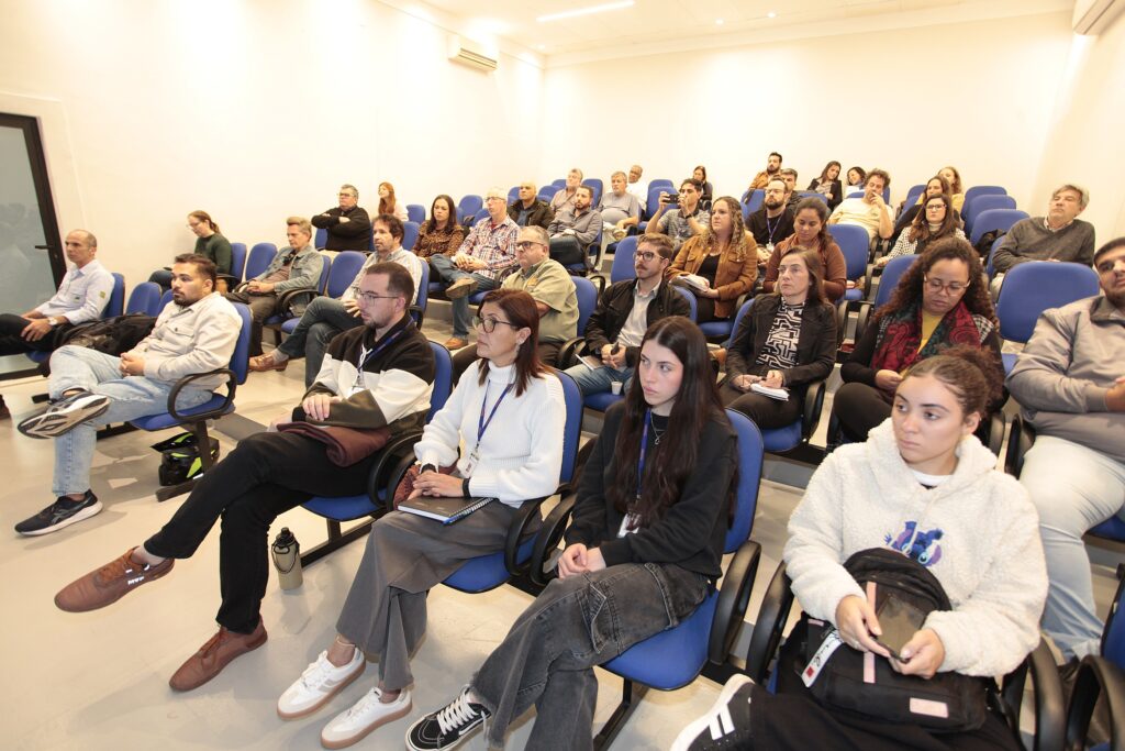 Esta foto mostra um auditório ou sala de treinamento preenchida por um público atento. • Composição: Vemos diversas fileiras de cadeiras azuis ocupadas por pessoas de diferentes idades, todas voltadas para o mesmo ponto, provavelmente acompanhando uma apresentação ou palestra. • Primeiro plano: Em destaque, na parte da frente, há um grupo diversificado de pessoas, incluindo jovens, homens e mulheres, vestindo roupas casuais. Vários deles têm cadernos ou mochilas por perto. • Ambiente: O espaço é bem iluminado, com paredes claras e teto liso, passando um aspecto profissional e funcional. O clima parece ser de aprendizado ou de uma reunião informativa.