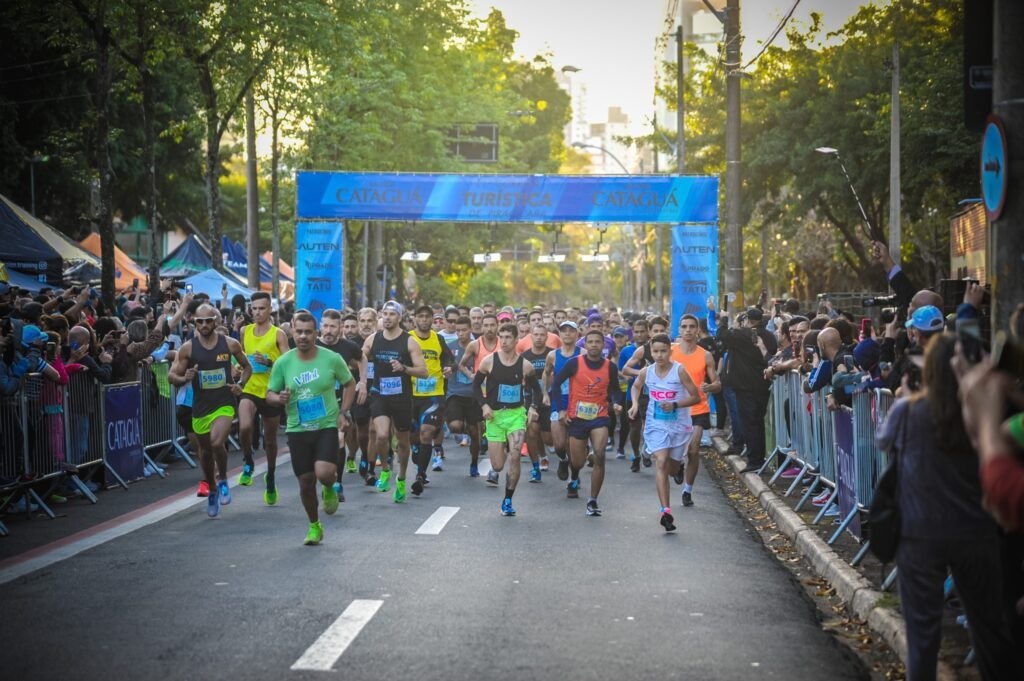 Esta foto captura o momento da largada de uma corrida de rua. Vemos um grande grupo de corredores vindo em direção à câmera, atravessando um pórtico azul de largada. Ambiente: A prova parece acontecer em uma rua arborizada durante o dia. Há grades de proteção nas laterais, com espectadores observando e tirando fotos dos participantes. Os Corredores: O pelotão da frente é composto principalmente por homens de diversas idades, todos vestindo roupas esportivas típicas (regatas, shorts e tênis de corrida) e usando números de peito. Eles parecem concentrados e em pleno movimento. Destaques: O clima é de energia e foco. No topo do pórtico, lê-se "Cataguá" e "Turística", indicando o nome do evento ou patrocinador. A iluminação é suave, sugerindo o início da manhã.