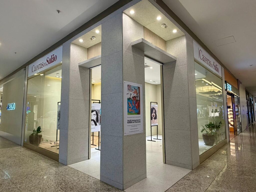 A imagem mostra a fachada de uma loja em um shopping, que está sendo usada como espaço para uma exposição cultural.Detalhes da cena:Loja: A fachada é da "Caipiras de Salão", com vitrines de vidro dos dois lados de um pilar central. O piso do corredor é de granito polido e brilhante, típico de shopping.Exposição: No pilar central tem um cartaz do "SALÃO Internacional de HUMOR de Piracicaba". O cartaz é colorido e mostra uma ilustração com personagens. Interior: Através das entradas abertas dá pra ver dentro da loja os mesmos totens pretos da foto anterior. Tem painéis com caricaturas em exposição, como o retrato do Paulo Leminski à esquerda e outro retrato à direita.Ambiente: Iluminação moderna com spots no teto. Ao lado, dá pra ver parte da fachada da loja "AREZZO" com letreiro azul neon. O corredor está vazio, sem circulação de pessoas no momento.A foto mostra como espaços comerciais foram adaptados para receber a mostra do Salão de Humor, levando arte e cultura para dentro do shopping e aproximando do público que circula pelo local.