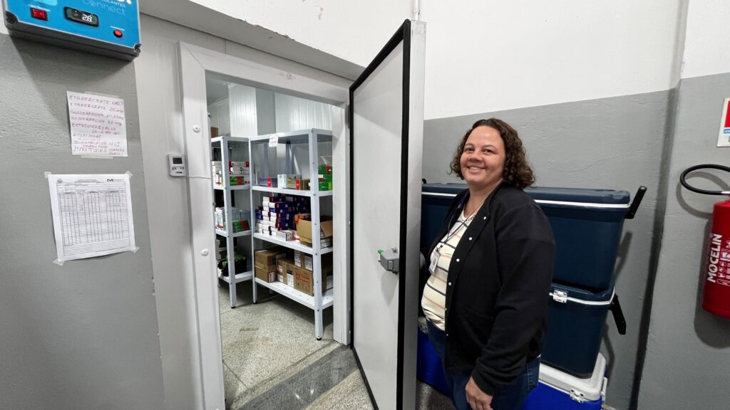 A imagem mostra uma mulher sorridente, de cabelos cacheados e castanhos, vestindo uma camiseta listrada sob um casaco preto, posando ao lado de uma câmara fria ou sala de armazenamento refrigerada com a porta aberta. Ao fundo, dentro da câmara, há prateleiras metálicas brancas organizadas com diversos medicamentos e caixas. Na parede ao lado da porta, há uma tela digital indicando a temperatura e uma lista manuscrita fixada, possivelmente contendo nomes de medicamentos armazenados, como "ETANERCEPTE", "GOSSERRELINA" e "RITUXIMABE". Na área à direita da mulher, há caixas térmicas azuis grandes e um extintor de incêndio vermelho preso à parede. O ambiente é limpo e parece ser um local controlado para armazenamento seguro de medicamentos, provavelmente em um hospital, clínica ou farmácia.