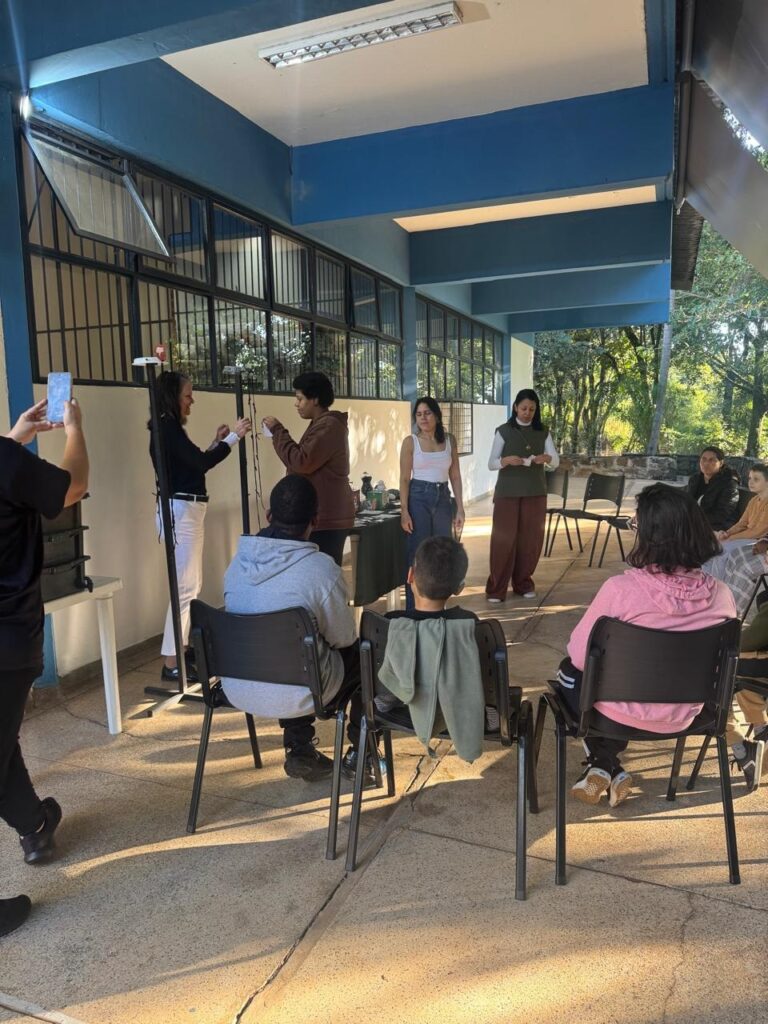 A imagem mostra um ambiente que parece ser um corredor ou uma área de passagem em um prédio, possivelmente uma escola ou centro comunitário. À esquerda, uma pessoa está tirando uma foto com um celular. No centro, duas mulheres estão de pé, aparentemente conduzindo uma atividade ou apresentação, ao lado de um suporte alto, que pode ser um tripé ou equipamento similar. À direita, várias pessoas, incluindo crianças e adultos, estão sentadas em cadeiras pretas, assistindo atentamente à apresentação. O local tem uma estrutura com pilares e vigas pintados de azul, e a iluminação natural entra pela lateral direita, criando sombras suaves no chão de concreto. A atmosfera parece ser calma e educativa, com um grupo reunido para uma atividade em conjunto.