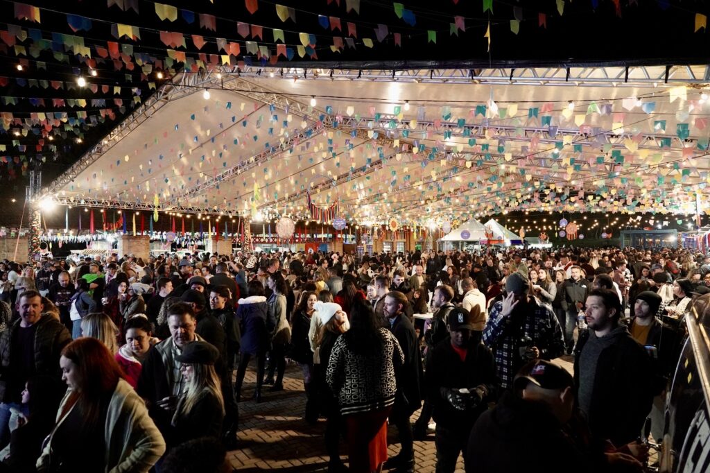 A imagem mostra uma festa ao ar livre durante a noite, provavelmente uma Festa Junina, devido às tradicionais bandeirinhas coloridas que estão penduradas em fileiras sob uma grande tenda. O ambiente está iluminado por luzes decorativas que criam uma atmosfera acolhedora e festiva. Há uma grande quantidade de pessoas reunidas, muitas delas vestindo roupas quentes como jaquetas, moletom e gorros, indicando que a temperatura está fria. As pessoas estão aglomeradas, conversando e aproveitando o evento. A cena transmite uma sensação de comunidade e celebração em um espaço público pavimentado. A foto foi tirada de um ponto um pouco elevado, permitindo uma visão ampla do evento e da multidão.