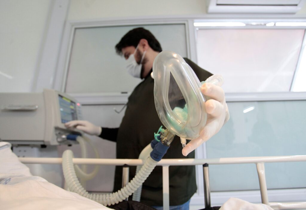 A imagem captura um profissional de saúde em ambiente hospitalar, segurando uma máscara de oxigênio e operando um equipamento médico ao lado de uma cama. A composição da cena é dinâmica, com o foco principal recaindo sobre a máscara de oxigênio em primeiro plano, que ocupa uma parte significativa do quadro, sugerindo uma ação iminente ou recente relacionada a um paciente. O profissional de saúde, desfocado em segundo plano, interage com um aparelho médico, indicando a continuidade dos cuidados. A cama hospitalar na parte inferior da imagem ancora a cena no contexto médico. A perspectiva é baixa, olhando para cima, o que confere uma certa dramaticidade ao ambiente. O principal sujeito visível é um profissional de saúde, com barba e cabelo escuros, vestindo uma máscara facial branca, luvas e um avental ou uniforme escuro. Sua expressão facial não é totalmente visível devido à máscara, mas a postura e a ação de mexer no aparelho sugerem atenção e profissionalismo. Ele está ajustando um equipamento médico, cujo painel exibe gráficos e números. A máscara de oxigênio, o outro elemento central, é transparente e de plástico, com conexões para tubos, e está posicionada de forma proeminente, com a mão enluvada do profissional segurando-a. A imagem parece ser uma fotografia, capturada com uma lente que proporciona uma profundidade de campo rasa, desfocando o fundo para destacar os elementos em primeiro plano e o profissional. O estilo é realista e documental, sem ornamentos artísticos, focando na clareza e na representação da situação médica. A iluminação é funcional, típica de um ambiente hospitalar, com luzes claras vindas de cima, sem sombras dramáticas. O cenário é claramente um quarto de hospital ou unidade de terapia intensiva. Ao fundo, observa-se uma janela com persianas ou divisórias de vidro, e um aparelho de ar condicionado no alto, elementos que definem o ambiente clínico. A cama hospitalar, com lençóis brancos, confirma o local. A iluminação geral é artificial e brilhante, mas não dura, proporcionando visibilidade adequada para os procedimentos médicos. A atmosfera geral é de cuidado médico, atenção e, possivelmente, de urgência ou recuperação.