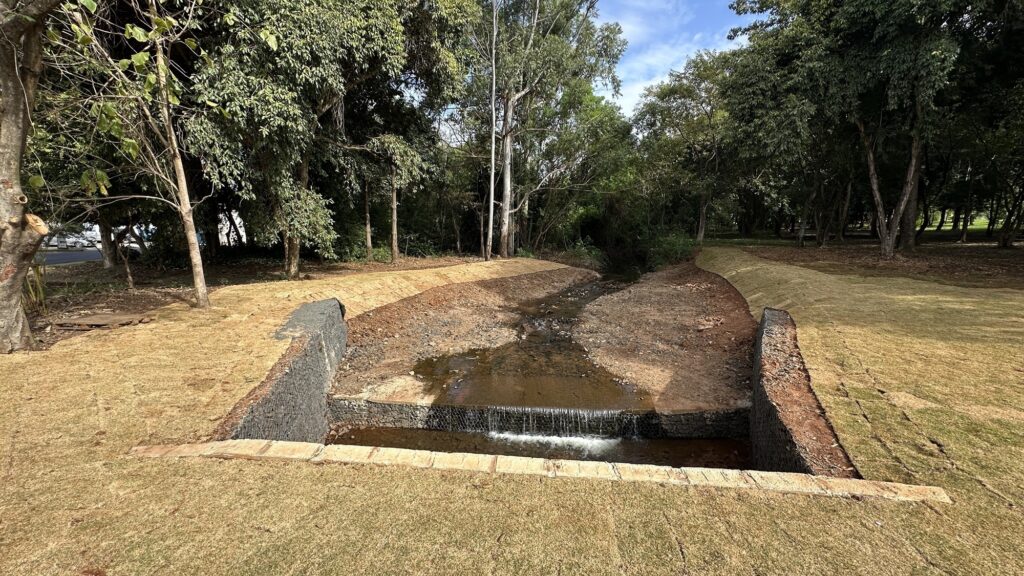A imagem mostra um pequeno córrego ou canal de drenagem em um ambiente de parque ou área verde. As margens do canal são reforçadas com gabiões, que são estruturas de arame preenchidas com pedras, formando paredes que contêm a água. A água desce formando uma pequena cascata sobre um desnível criado pelos gabiões. Ao redor do canal, há um gramado recém-plantado, que ainda parece um pouco seco ou amarelado. Ao fundo, observa-se uma área com árvores maduras, criando um cenário natural e tranquilo. O céu visível entre as árvores indica um dia claro ou parcialmente ensolarado. A imagem transmite uma sensação de integração entre a engenharia para controle da água e o ambiente natural ao redor.