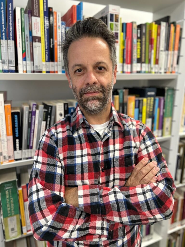 A imagem mostra um homem de meia-idade, com cabelo curto e grisalho, e barba aparada. Ele está em pé, com os braços cruzados, usando uma camisa xadrez em tons de vermelho, branco, azul e preto, e uma camiseta branca por baixo. O fundo é uma estante cheia de livros organizados, alguns com títulos visíveis como "Língua Brasileira de Sinais", "Cambridge", "Carl Sagan", "Cosmos", "Estatística" e "História da Informação". A iluminação é suave, destacando o homem no centro da imagem, enquanto os livros ao fundo ficam ligeiramente desfocados, criando um ambiente que sugere conhecimento, estudo ou ambiente acadêmico.