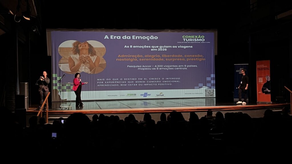 Nesta imagem, vemos uma apresentação em um auditório sobre "A Era da Emoção", focada em tendências de turismo para 2026. No telão, há informações sobre uma pesquisa da Accor com 4.300 viajantes, listando as 8 emoções que guiam as viagens atualmente: admiração, alegria, liberdade, conexão, nostalgia, serenidade, surpresa e prestígio. No palco: • À esquerda, uma intérprete de Libras faz a tradução simultânea. • Ao centro, uma palestrante com um blazer rosa fala ao público. • À direita, um homem está em pé próximo a um banner. Há também o logo "Conexão Turismo" e os logotipos dos realizadores (Prefeitura de Piraí, Sebrae e Senac) na parte inferior da projeção.