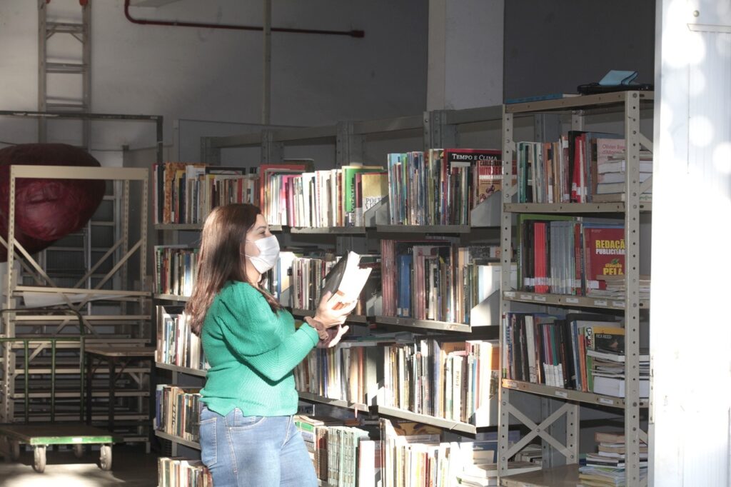 A imagem mostra uma mulher em uma biblioteca ou arquivo, vista de perfil enquanto escolhe um livro em uma estante. Ela tem cabelos castanhos longos e usa uma blusa verde vibrante, calça jeans azul e uma máscara branca no rosto. A mulher está segurando um livro aberto, aparentemente lendo ou examinando seu conteúdo. Ao redor, há várias estantes metálicas repletas de livros de diferentes tamanhos e cores. A iluminação é natural, entrando provavelmente por uma janela próxima, criando sombras e realces que destacam a cena. À esquerda da imagem, há um carrinho metálico com um objeto vermelho de formato abstrato apoiado sobre ele. Os títulos visíveis em alguns livros incluem "TEMPO E CLIMA" e "REDAÇÃO PUBLICITÁRIA". O ambiente parece funcional, com estrutura exposta e uma escada ao fundo, reforçando o clima de espaço dedicado ao estudo ou pesquisa.