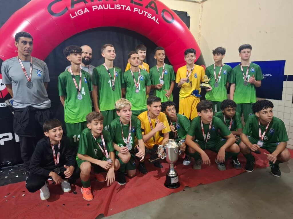 A imagem mostra um time de futsal juvenil posando para uma foto de comemoração, provavelmente após uma conquista. • O cenário: Eles estão em frente a um arco inflável vermelho da "Liga Paulista Futsal". Há um tapete vermelho no chão e, no centro, um troféu grande e prateado. • O time: Há um grupo de jovens jogadores vestindo uniformes predominantemente verdes, com alguns vestindo amarelos. Quase todos usam medalhas no pescoço. • Detalhes: Um adulto, que parece ser o treinador ou um membro da comissão técnica, está de pé no lado esquerdo, usando uma camisa cinza. Os jogadores estão dispostos entre fileiras em pé e agachados, sorrindo e celebrando o momento.