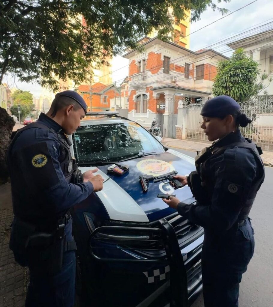 A imagem mostra dois policiais em uma rua urbana durante o dia. Eles estão posicionados em ambos os lados do capô de uma viatura policial, que é azul com detalhes brancos e tem um emblema visível na frente. O policial à esquerda é um homem, vestido com uniforme azul escuro, colete tático e boina, olhando atentamente para algo em suas mãos. A policial à direita é uma mulher, também com uniforme azul escuro e boina, segurando uma arma de fogo e um carregador. Sobre o capô do carro, há duas pistolas, um carregador e um pequeno dispositivo eletrônico. Ao fundo, há uma casa grande de vários andares, com fachada em tons claros e janelas grandes. A cena parece ser uma inspeção ou preparação de armamento, transmitindo um ambiente profissional e sério. A palavra "POLICIA" está visível na viatura, indicando que se trata da polícia local.