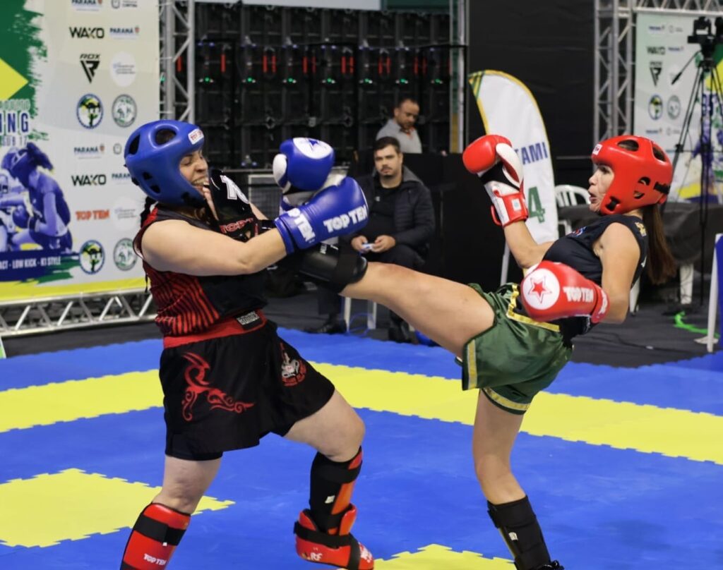 A imagem mostra uma luta de kickboxing entre duas atletas femininas em um ringue com piso azul e amarelo. A lutadora à esquerda está vestindo capacete azul, luvas azuis, camiseta preta sem mangas com detalhes vermelhos, shorts pretos com desenhos vermelhos e protetores de canela. Ela está bloqueando um chute alto da adversária. A lutadora à direita usa capacete vermelho, luvas vermelhas, camiseta preta e shorts verdes com detalhes dourados, também com protetores de canela, e está executando um chute alto na direção da oponente. Ao fundo, há banners com as marcas "WAKO", "PARANA", "TOP TEN" e "RDX", indicando que o evento é oficial e possivelmente uma competição de alto nível. Também é possível ver uma pessoa sentada, provavelmente um árbitro ou treinador, observando a luta. A cena transmite a intensidade e o dinamismo do esporte.