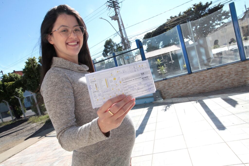 A imagem mostra uma mulher grávida sorridente, usando óculos e um suéter cinza claro, segurando um comprovante de vacinação em direção à câmera. Ela está ao ar livre, em um local ensolarado, com árvores e um muro baixo ao fundo. O comprovante é da Secretaria de Estado da Saúde de São Paulo, indicando que a vacina aplicada foi contra a Influenza, com a data 23/03/16. A mulher parece feliz e orgulhosa ao mostrar o documento. A luz do sol ilumina a cena, criando um ambiente claro e agradável.