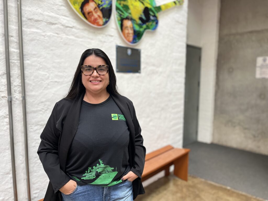 Nesta foto, uma mulher de cabelos pretos e longos, sorridente, está posando para a câmera. Ela usa óculos com armação estampada, uma camiseta preta com uma estampa verde na parte inferior (que parece ser um navio) e o logo "Ponta Verde", além de um blazer preto aberto por cima. Ela está em um ambiente interno com paredes brancas, onde há uma decoração colorida na parede (parecendo guitarras decoradas) acima de uma placa informativa. Atrás dela, nota-se um banco de madeira e uma porta à direita.