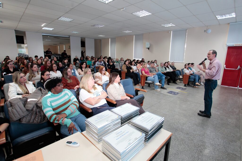 A imagem mostra um evento ou palestra acontecendo em um auditório ou sala de conferências. Um homem está de pé à direita, segurando um microfone e falando para um grupo majoritariamente composto por mulheres, que estão sentadas em poltronas azuis, ouvindo atentamente. Em primeiro plano, sobre uma mesa, há várias pilhas de certificados ou diplomas encadernados, prontos para serem entregues. O ambiente parece ser bem iluminado e funcional, com paredes em tons claros, piso cinza e uma porta de emergência vermelha ao fundo. O clima parece ser de uma cerimônia, talvez de entrega de certificados ou uma formação acadêmica.