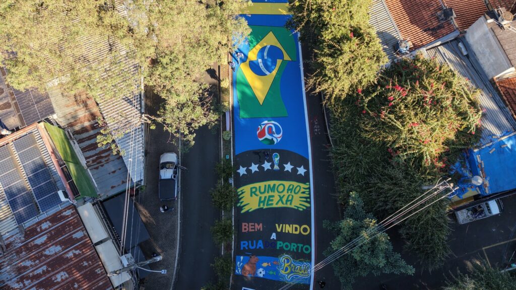 A imagem mostra uma rua vista de cima, com uma pintura colorida e vibrante no asfalto que celebra o futebol brasileiro e a Copa do Mundo da FIFA. No centro da pintura, há a bandeira do Brasil com as palavras "ORDEM E PROGRESSO", além de uma bola de futebol estilizada e o troféu da Copa do Mundo. Acima da bandeira, está escrito "RUMO AO HEXA", indicando a esperança de conquistar o sexto título mundial. Abaixo, há a frase "BEM VINDO A RUA DO PORTO". Também aparecem elementos gráficos como cinco estrelas, representando os títulos já conquistados pelo Brasil, e desenhos de animais típicos, como um capivara e peixes, junto com a palavra "Brasil". A rua está cercada por árvores verdes e algumas casas com telhados de metal e painéis solares. Um carro prata está estacionado à esquerda. A iluminação sugere que a foto foi tirada durante o dia, com sombras longas projetadas pelas árvores. A pintura é detalhada e ocupa uma grande parte da rua, demonstrando uma celebração cultural e esportiva local.