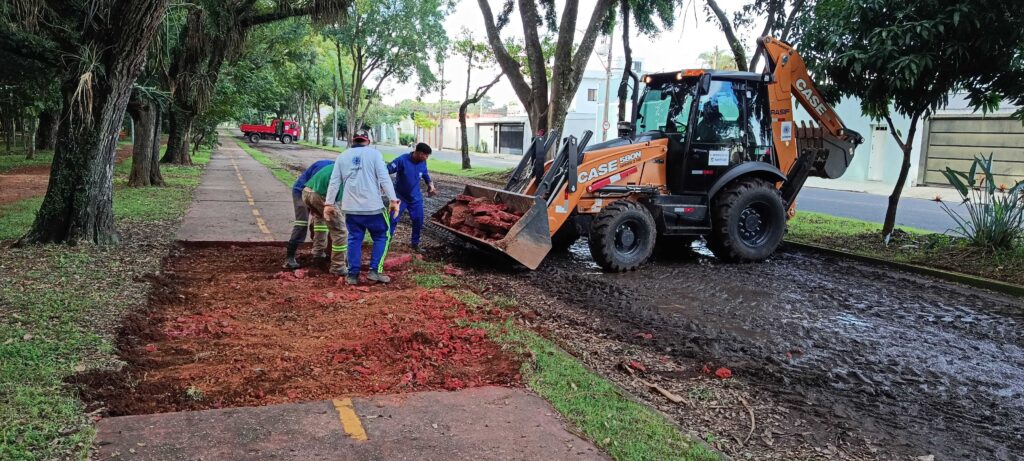A imagem mostra uma equipe de trabalhadores realizando uma obra de manutenção em uma via pública, possivelmente uma ciclovia ou calçada, em uma área arborizada. * Em primeiro plano, quatro homens uniformizados estão posicionados próximos a uma retroescavadeira da marca CASE (modelo 580N). Eles parecem estar auxiliando na remoção ou organização de pedaços de concreto e entulho avermelhado que estão sendo recolhidos pela caçamba frontal da máquina. * O terreno ao redor da máquina está bastante enlameado, sugerindo condições climáticas recentes de chuva ou um solo úmido devido à escavação. * Ao fundo, é possível observar uma rua asfaltada com casas, além de um caminhão basculante vermelho estacionado mais adiante na via, indicando que o local está sendo preparado para o transporte de materiais. * O ambiente é cercado por árvores grandes, o que confere um aspecto de parque ou via urbana arborizada. A cena transmite uma atividade típica de infraestrutura municipal, com a presença de sinalização e equipamentos pesados para a movimentação de terra e detritos.