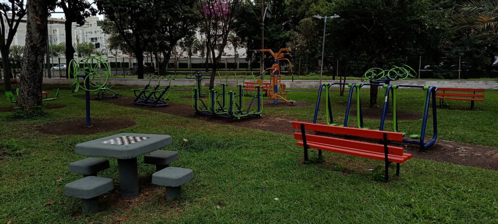 A imagem mostra um parque público ao ar livre, com várias áreas destinadas ao lazer e exercícios físicos. Em primeiro plano, há uma mesa de concreto para jogar xadrez, acompanhada por quatro pequenos bancos também de concreto. Ao lado da mesa, há um banco vermelho colocado sobre a grama. Mais ao fundo, é possível ver diversos aparelhos de ginástica ao ar livre, em cores vibrantes como verde, azul e laranja, incluindo equipamentos para exercícios de braços e pernas. O parque está cercado por árvores e gramado bem cuidado, e ao fundo há prédios residenciais de vários andares. O céu está nublado, criando uma iluminação suave e difusa na cena. O ambiente transmite uma sensação de espaço aberto e convidativo para atividades físicas e recreação.