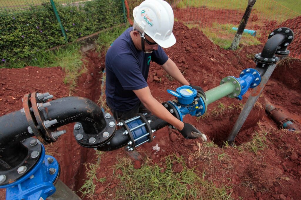 A imagem mostra um trabalhador da construção civil, usando capacete branco com o logo "SANE SOLUTI" e camiseta azul escura, realizando um serviço em uma tubulação de água. Ele está agachado dentro de uma vala aberta no solo de terra vermelha, utilizando uma ferramenta para apertar ou ajustar uma conexão na tubulação. A tubulação é composta por tubos de metal e plástico, com partes azuis e verdes, além de conexões e parafusos visíveis. Ao fundo, há uma cerca de segurança laranja e vegetação verde ao redor da vala. A cena transmite um ambiente de trabalho ao ar livre, provavelmente em uma obra de saneamento ou infraestrutura hidráulica.