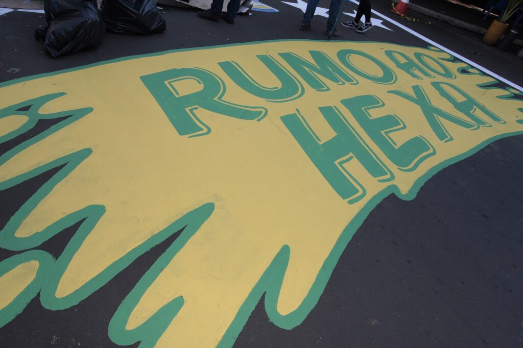 A imagem mostra uma pintura grande no chão de uma rua asfaltada. A arte é feita com tinta amarela e contornos verdes, formando uma figura que lembra um respingo ou uma mancha irregular. Dentro dessa figura está escrito, em letras maiúsculas e grandes, "RUMO AO HEXA". Esse é um slogan popular no Brasil, usado para expressar a expectativa e o apoio à seleção brasileira de futebol na busca pelo sexto título mundial (o "hexa" refere-se ao sexto campeonato da Copa do Mundo). Ao redor da pintura, é possível ver os pés de algumas pessoas que estão próximas, sacos de lixo pretos à esquerda e um cone de trânsito junto com um vaso de planta à direita. A foto foi tirada de um ângulo elevado, mostrando a arte no chão em perspectiva.