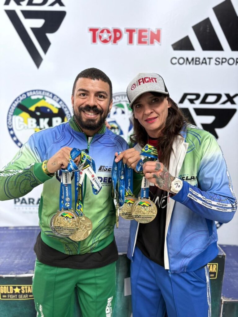 A imagem mostra dois atletas, um homem e uma mulher, posando juntos em um pódio ou local de premiação, ambos sorrindo e exibindo várias medalhas de ouro penduradas no pescoço. Eles vestem roupas esportivas com cores que remetem ao Brasil (verde, azul e amarelo). A mulher usa um boné cinza com a palavra "FIGHT" e tem tatuagens visíveis nas mãos e braços. O homem tem barba curta e cabelo escuro. Ao fundo, há um banner branco com vários logotipos, incluindo o da Confederação Brasileira de Kickboxing (C.B.K.B.), além de marcas como RDX, TOP TEN e COMBAT SPORTS. A imagem transmite uma sensação de vitória e celebração no contexto do kickboxing brasileiro, provavelmente após uma competição nacional.