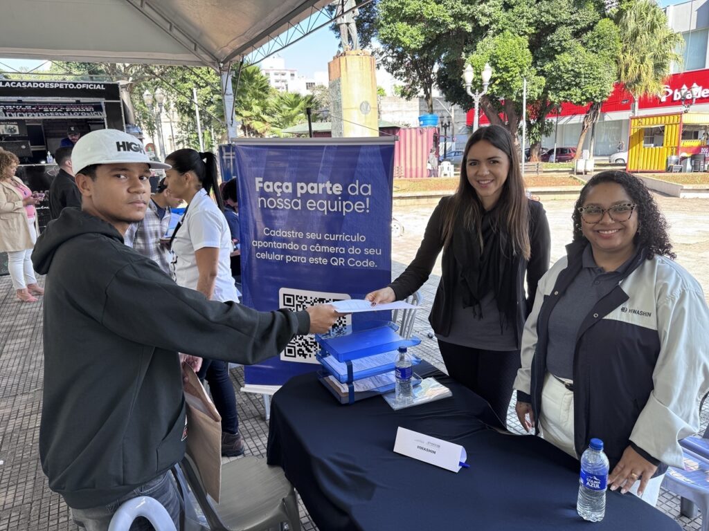 A imagem retrata uma ação de recrutamento ao ar livre em uma praça pública. Em primeiro plano, um jovem de boné branco e casaco escuro entrega um currículo em papel para uma mulher que está atrás de uma mesa coberta com um pano preto. Ao lado da mulher, outra pessoa sorri para a câmera, vestindo uma jaqueta com a inscrição "HWASHIN". Sobre a mesa, há uma placa com o nome da empresa, garrafas de água e uma bandeja de documentos. Ao fundo, destaca-se um banner azul com a frase "Faça parte da nossa equipe!" e um QR Code para cadastro de currículos online. O ambiente é iluminado pelo sol, com árvores, calçada e um monumento ao fundo, sugerindo um evento de empregabilidade em um espaço urbano.