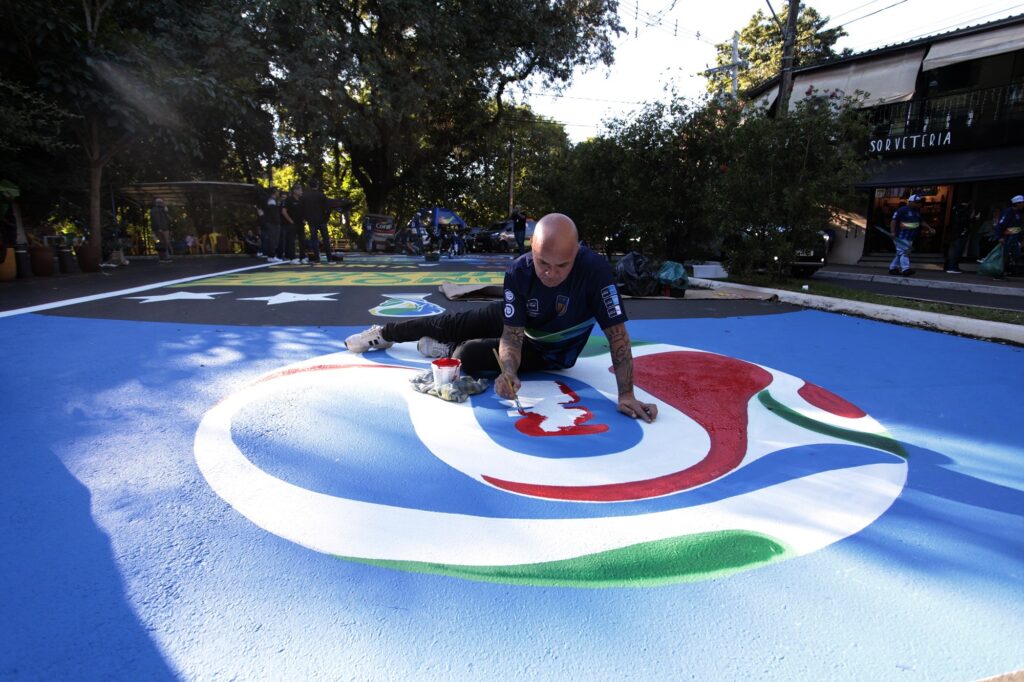 A imagem mostra um homem ajoelhado no asfalto de uma rua, pintando um grande mural colorido diretamente no chão. Ele é careca, tem tatuagens nos braços e veste uma camisa polo azul escura e calças pretas. O homem está concentrado enquanto pinta uma figura abstrata e estilizada com cores vibrantes, incluindo azul, branco, vermelho e verde. Ao lado dele, há um recipiente com tinta vermelha. Ao fundo, é possível ver árvores, algumas pessoas em pé e um prédio com uma placa que diz "SORVETERIA". A cena ocorre durante o dia, com luz natural e sombras visíveis no chão. A perspectiva da foto é levemente elevada, mostrando o artista e sua obra no chão.
