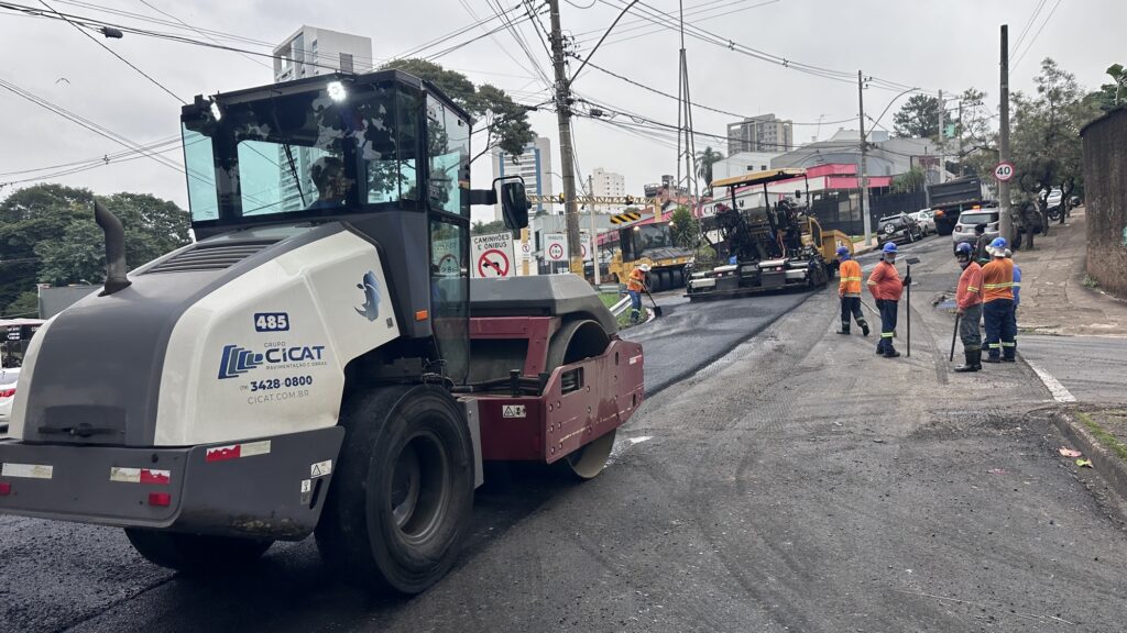 A imagem mostra uma cena de obras de pavimentação em uma rua urbana. Em primeiro plano, destaca-se um grande rolo compressor da empresa Grupo CICAT Pavimentação e Obras, identificado pelo número “485” e informações de contato. Ao redor, vários trabalhadores vestindo coletes laranja, capacetes azuis e calças refletivas atuam sobre o asfalto recém-aplicado. Mais ao fundo, outra máquina pesada espalha o material, enquanto placas de trânsito indicam restrições como “Caminhões e Ônibus” e o limite de velocidade de “40”. O cenário é complementado por prédios, fiação elétrica e veículos estacionados, compondo um retrato típico de manutenção de infraestrutura urbana, onde o trabalho coletivo garante melhorias na mobilidade e na qualidade das vias.