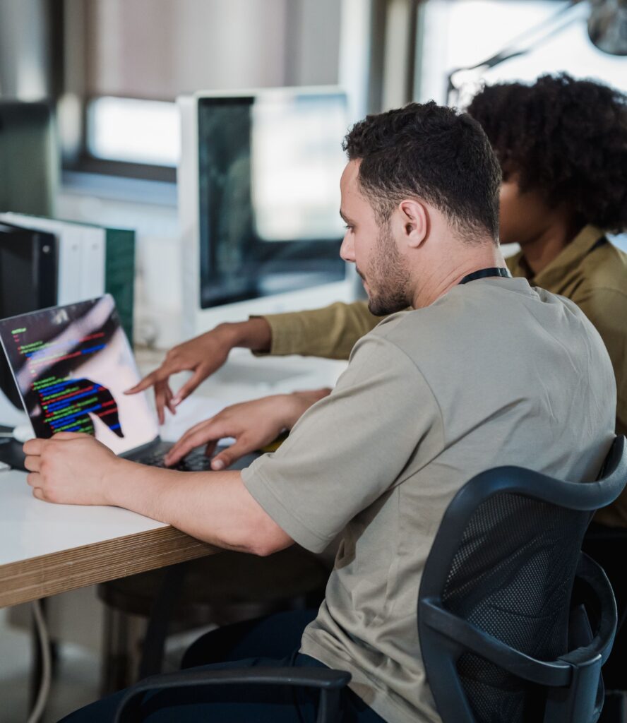 A imagem mostra uma cena de escritório onde duas pessoas estão concentradas em um laptop. Em primeiro plano, um homem de camiseta clara está sentado em uma cadeira de escritório preta, digitando no laptop. Atrás dele, uma mulher com cabelo cacheado e vestindo uma blusa mostarda aponta para a tela do laptop, sugerindo que está ajudando ou discutindo algo relacionado ao que está sendo exibido. A tela do laptop exibe linhas de código coloridas, indicando que eles estão trabalhando com programação. O ambiente é moderno, com uma mesa branca e um monitor de computador grande ao fundo, iluminado por uma luz suave e natural. A imagem transmite uma atmosfera de colaboração profissional entre os dois.
